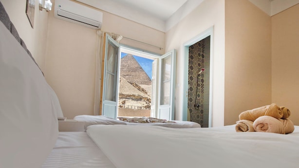 A hotel room with a bed, a window with a view of a pyramid, and a wall-mounted air conditioner. - Villa Khufu Pyramids Hotel (Giza)