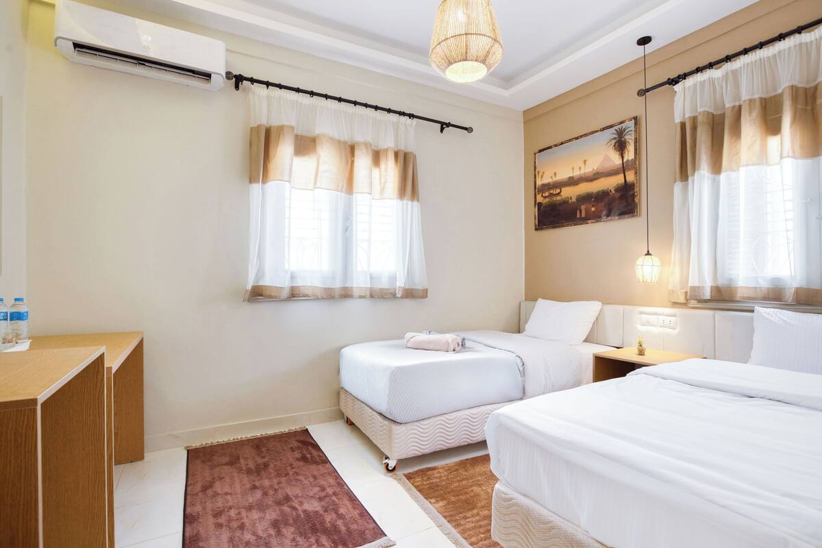 Une chambre d’hôtel avec deux lits, un bureau, un appareil de climatisation, un tableau et une fenêtre avec des rideaux.