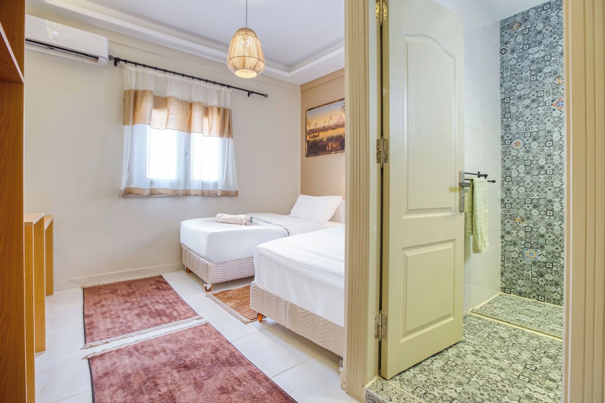 Un dormitorio con dos camas, una ventana con cortinas, una lámpara colgante y un baño con azulejos estampados.