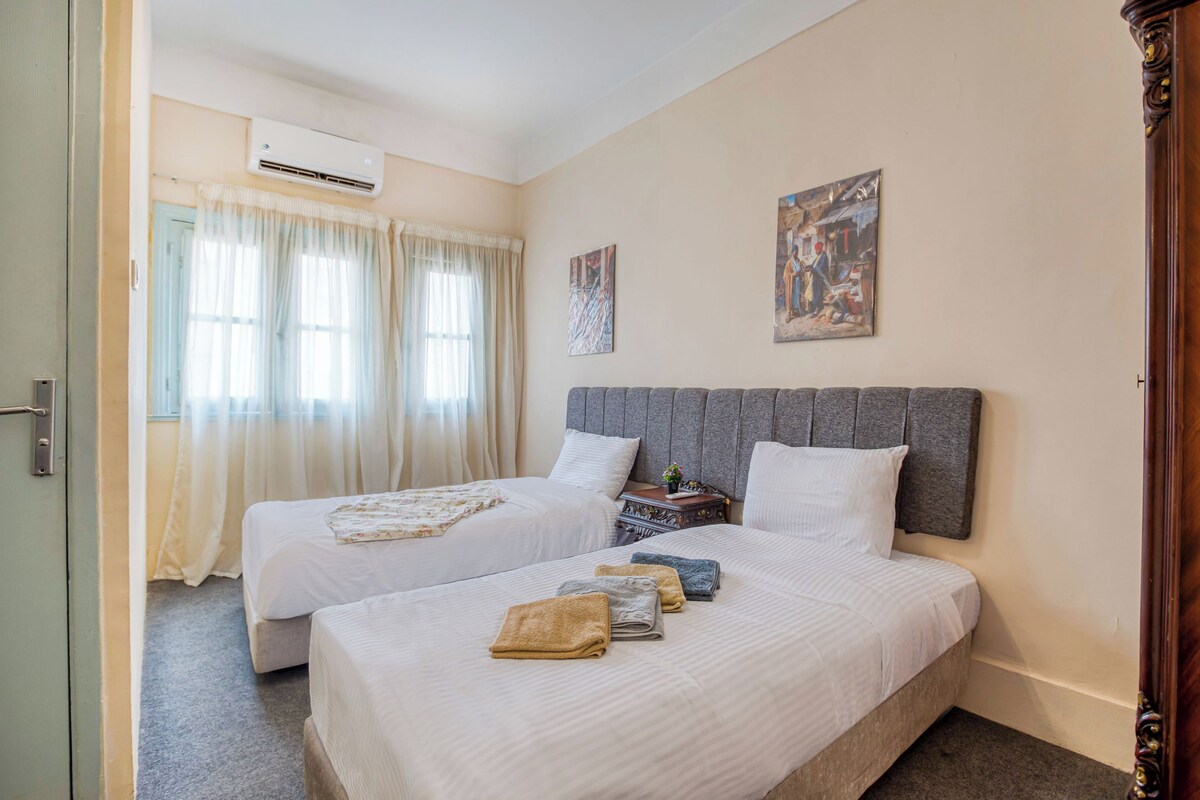 Habitació d'hotel amb dos llits, una tauleta de nit, un aparell d'aire condicionat i dos quadres a la paret.