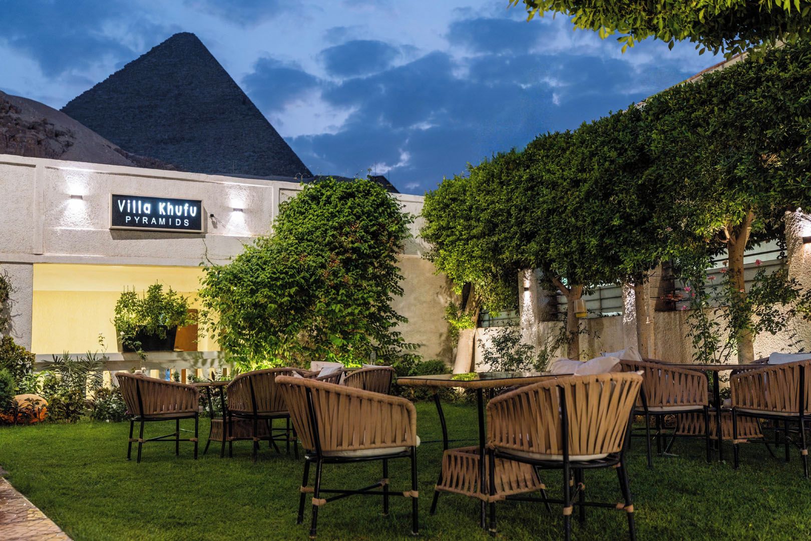 An outdoor seating area with wicker chairs and tables, a sign for Villa Khufu Pyramids, and a pyramid in the background.