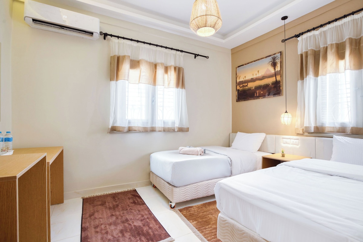 A hotel room with two beds, a desk, an air conditioning unit, a painting, and a window with curtains.