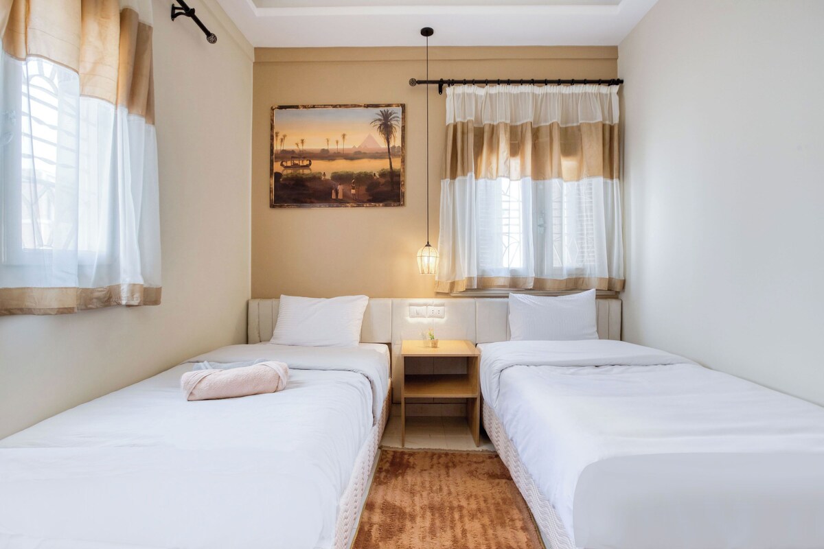 Una habitació d'hotel amb dos llits individuals, una tauleta de nit amb una làmpada, un quadre a la paret i una finestra amb cortines.
