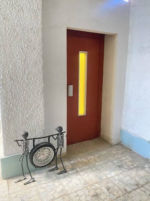 A hallway with a wheelchair, a door with a yellow light, and a textured wall.