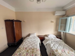 A room with two beds, a wooden wardrobe, a small table, and an air conditioning unit.