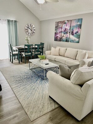 A living room with a beige sofa, a coffee table, a dining table with chairs, a clock, and abstract wall art.