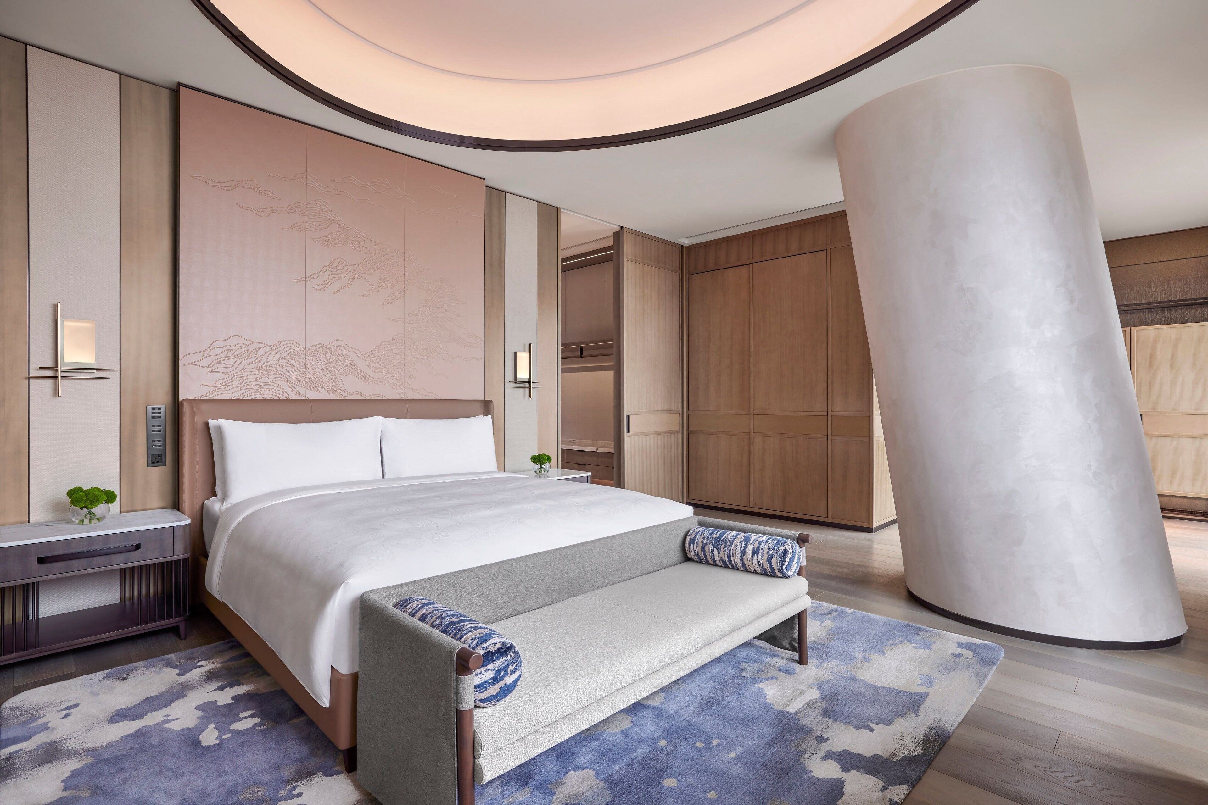 A modern hotel room with a large bed, a bench, a large cylindrical column, and a wall with a wave design.