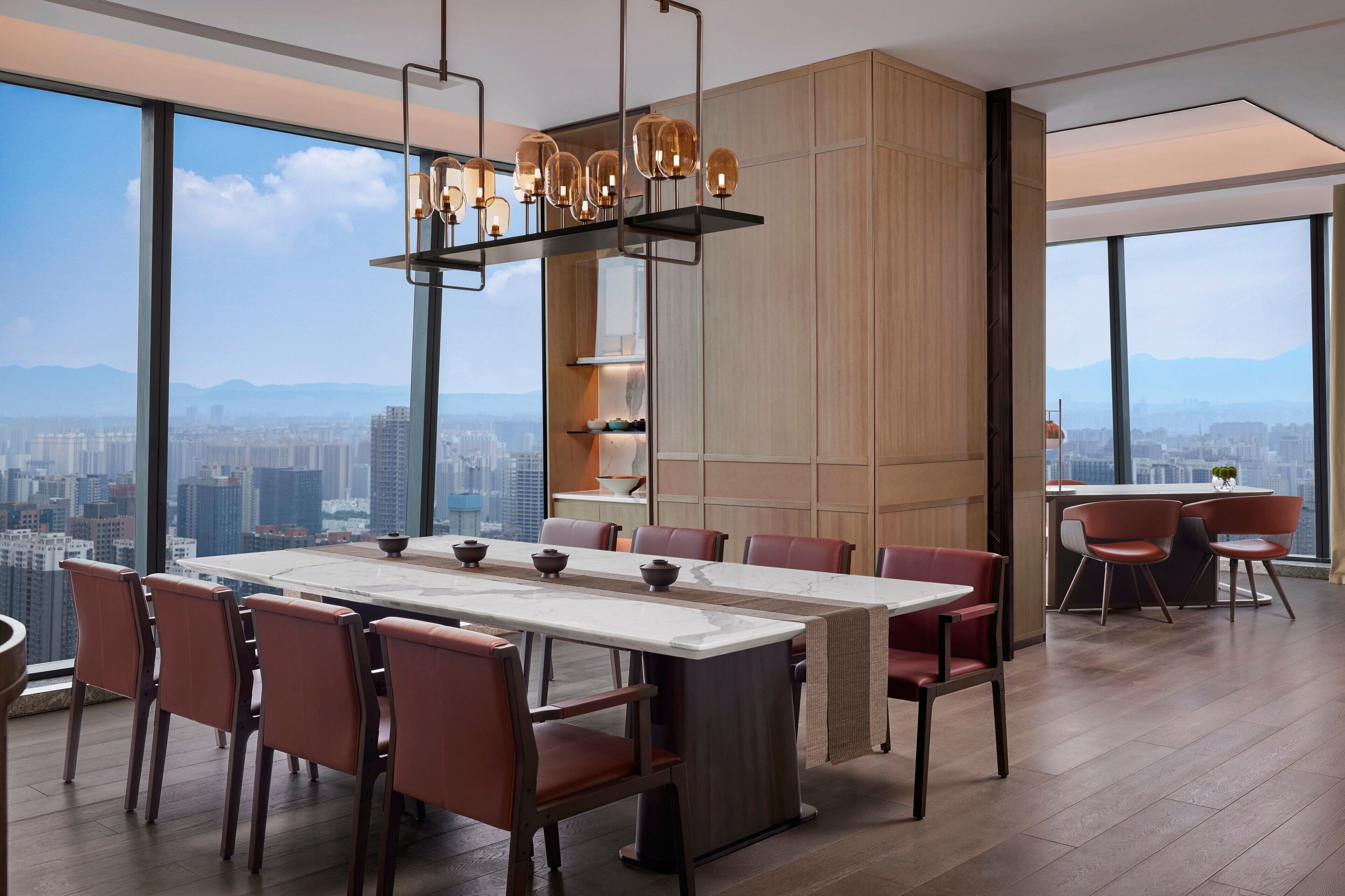 A modern dining area with a large table and chairs, a unique chandelier, and a city view through large windows.