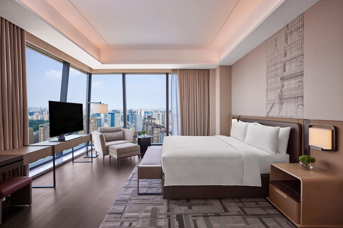 A modern hotel room with a large bed, a desk with a flat-screen TV, a comfortable armchair, a small table, and a city view through large windows.