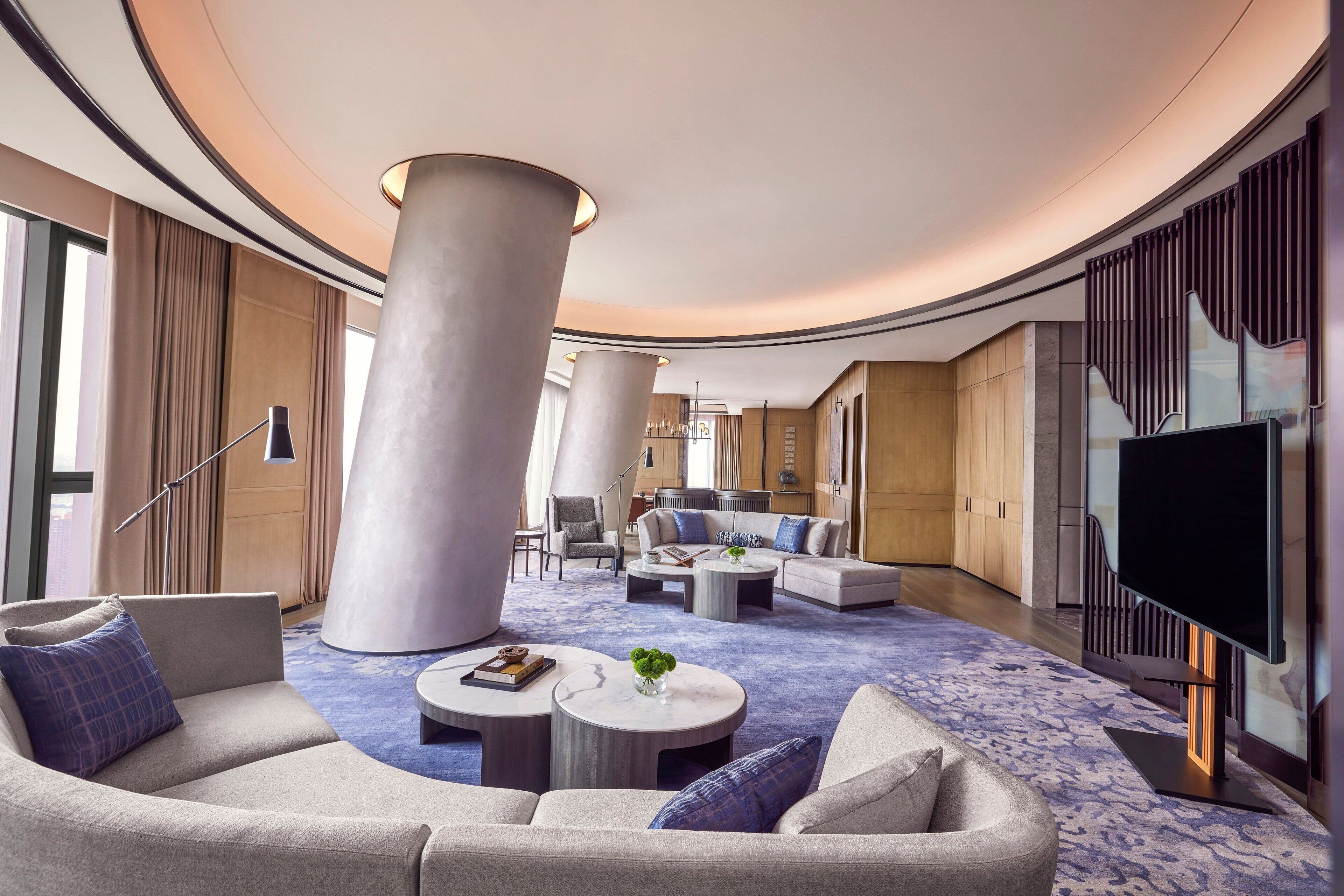 A modern hotel lobby with a curved ceiling, large columns, a flat-screen TV, and a seating area with sofas and round tables.
