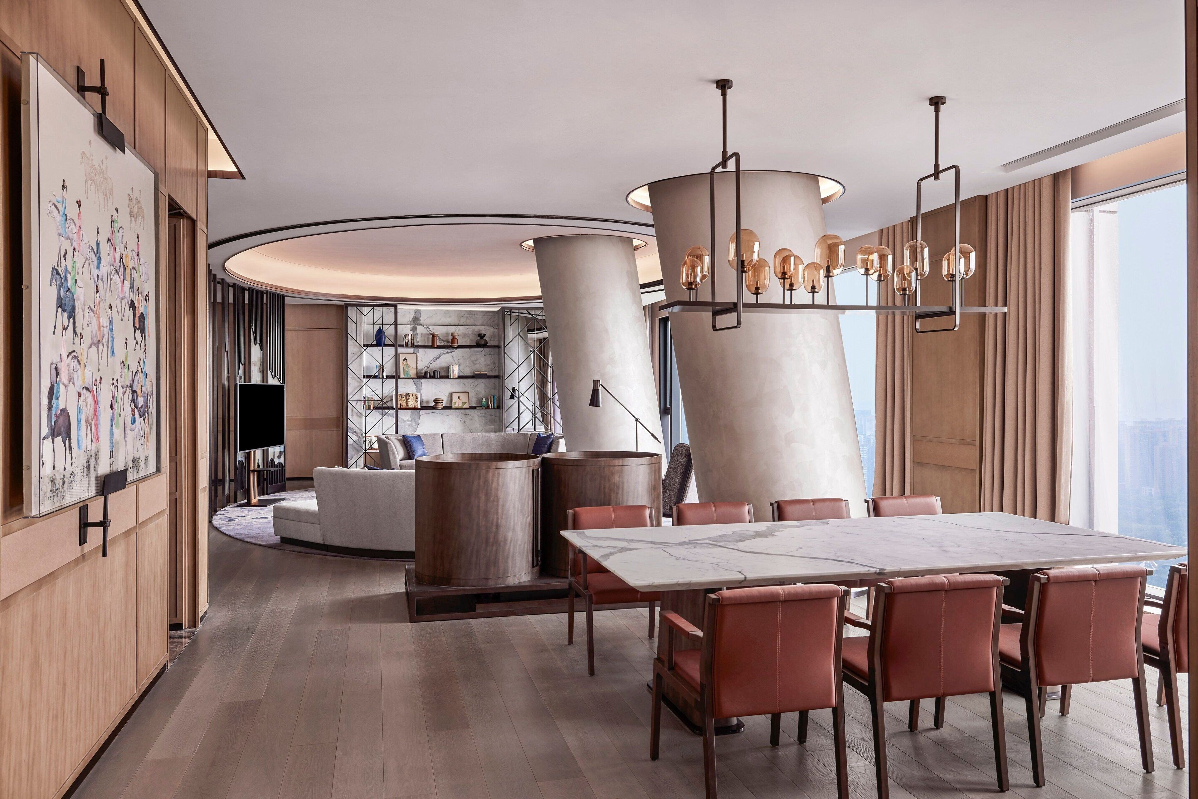 A modern hotel lobby with a large dining table, leather chairs, a unique chandelier, and a view of the city.