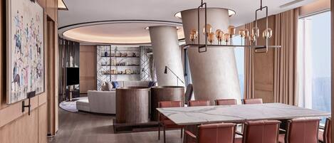 A modern hotel lobby with a large dining table, leather chairs, a unique chandelier, and a view of the city.