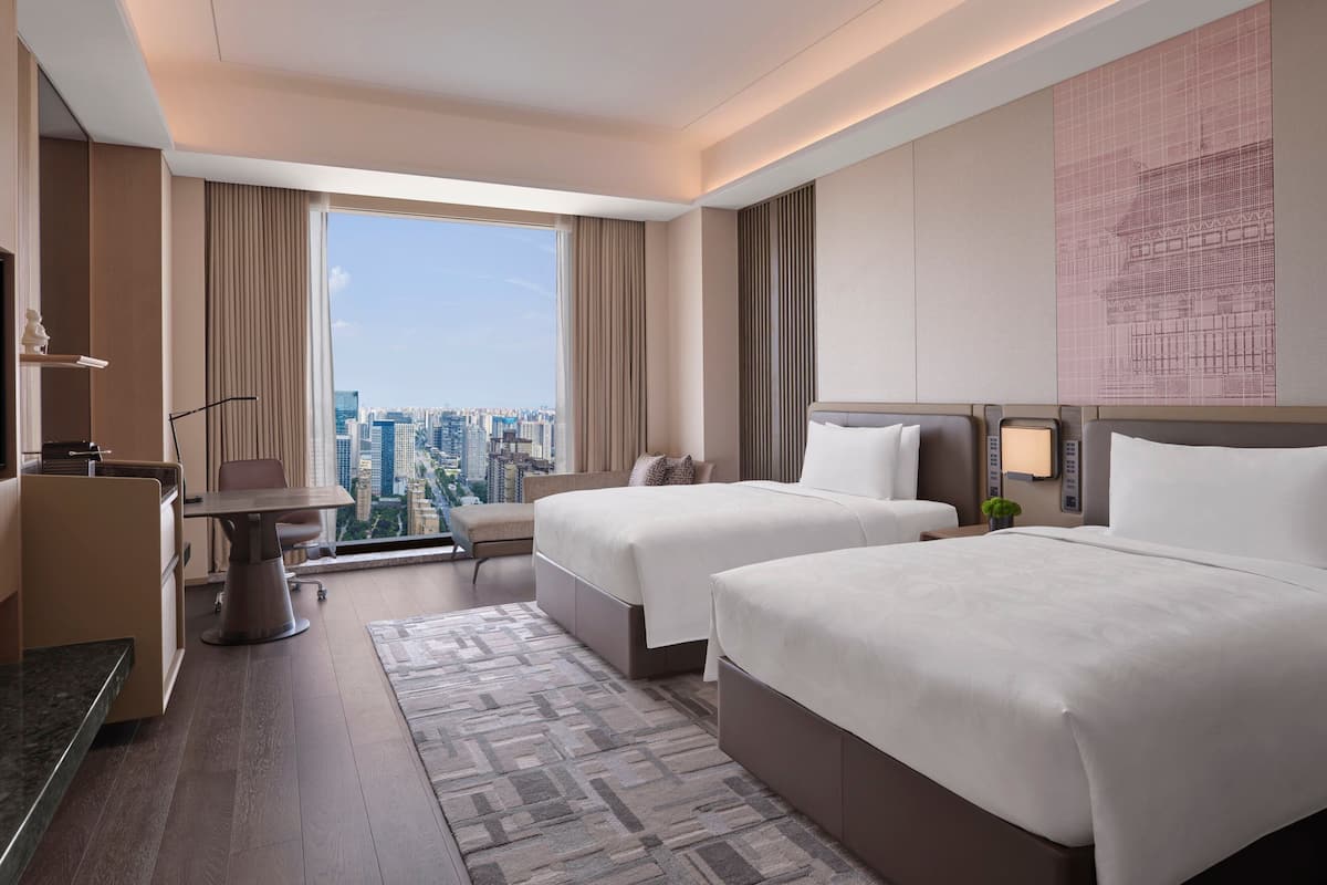A hotel room with two beds, a desk, a chair, a small table, and a large window offering a city view.