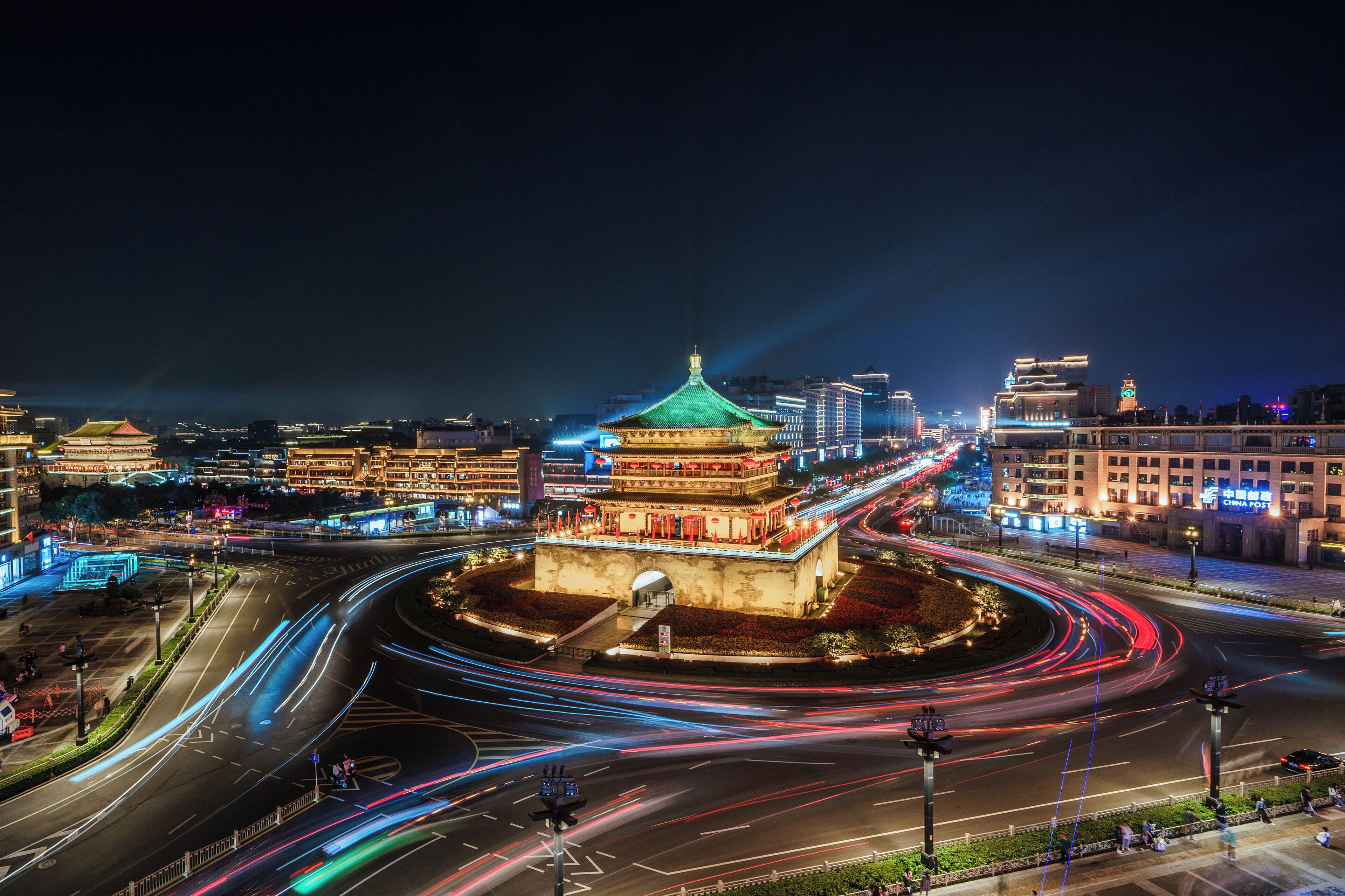 A historic building with a green roof and traditional architecture, surrounded by modern city lights and a busy intersection with light trails.