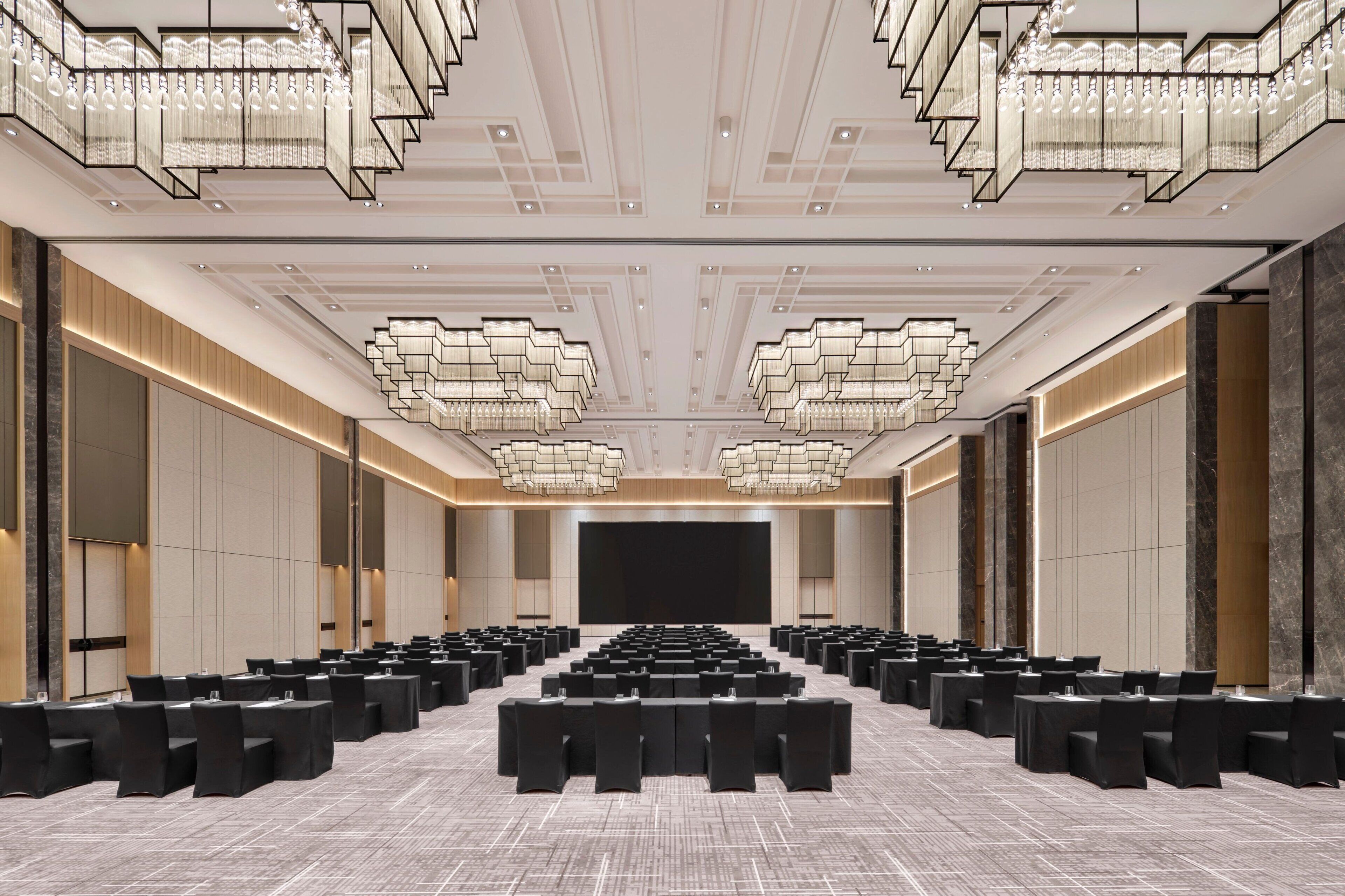 A conference room with rows of tables and chairs, a large screen at the front, and modern chandeliers.