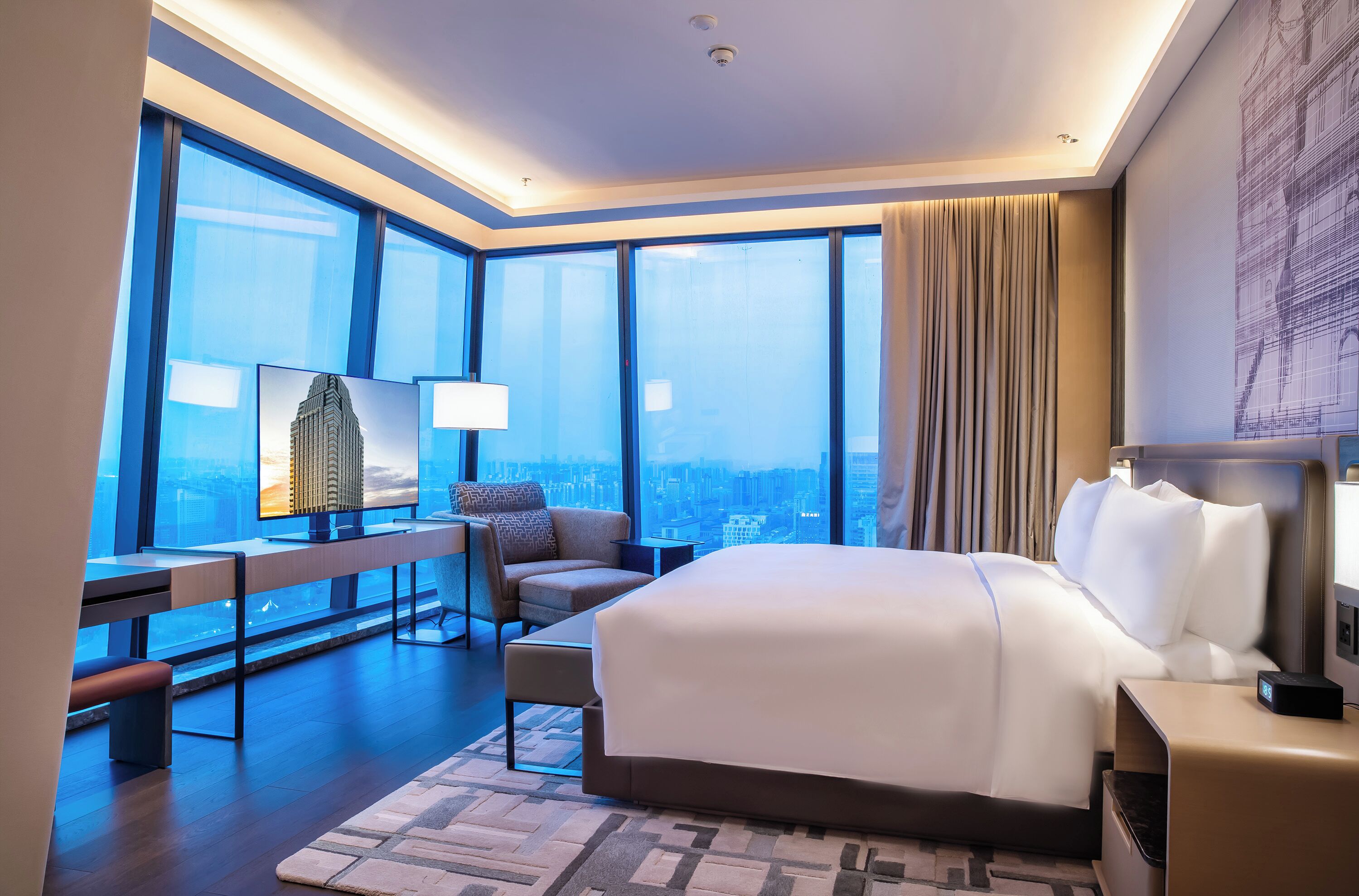 A modern hotel room with a large bed, a desk, a chair, and a view of the city through large windows.
