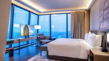 A modern hotel room with a large bed, a desk, a chair, and a view of the city through large windows.