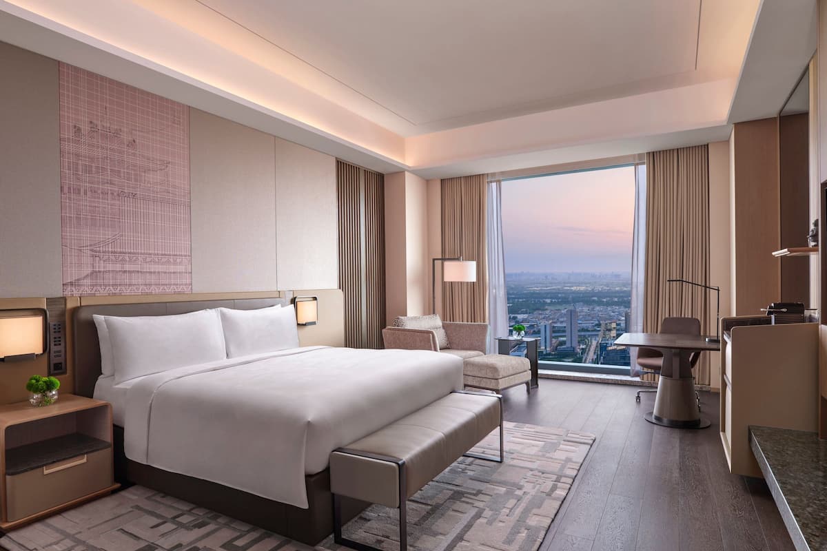 A modern hotel room with a large bed, a desk, a chair, and a view of the city.