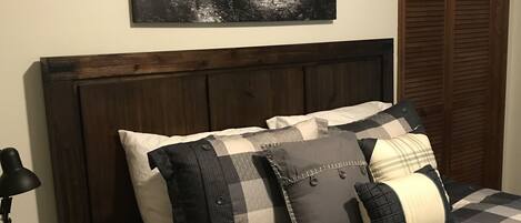 A bedroom with a dark wood headboard, a checkered bedspread, and a black and white forest-themed wall art.