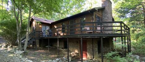A rustic cabin with a wooden deck, a stone chimney, and a gravel driveway surrounded by trees.