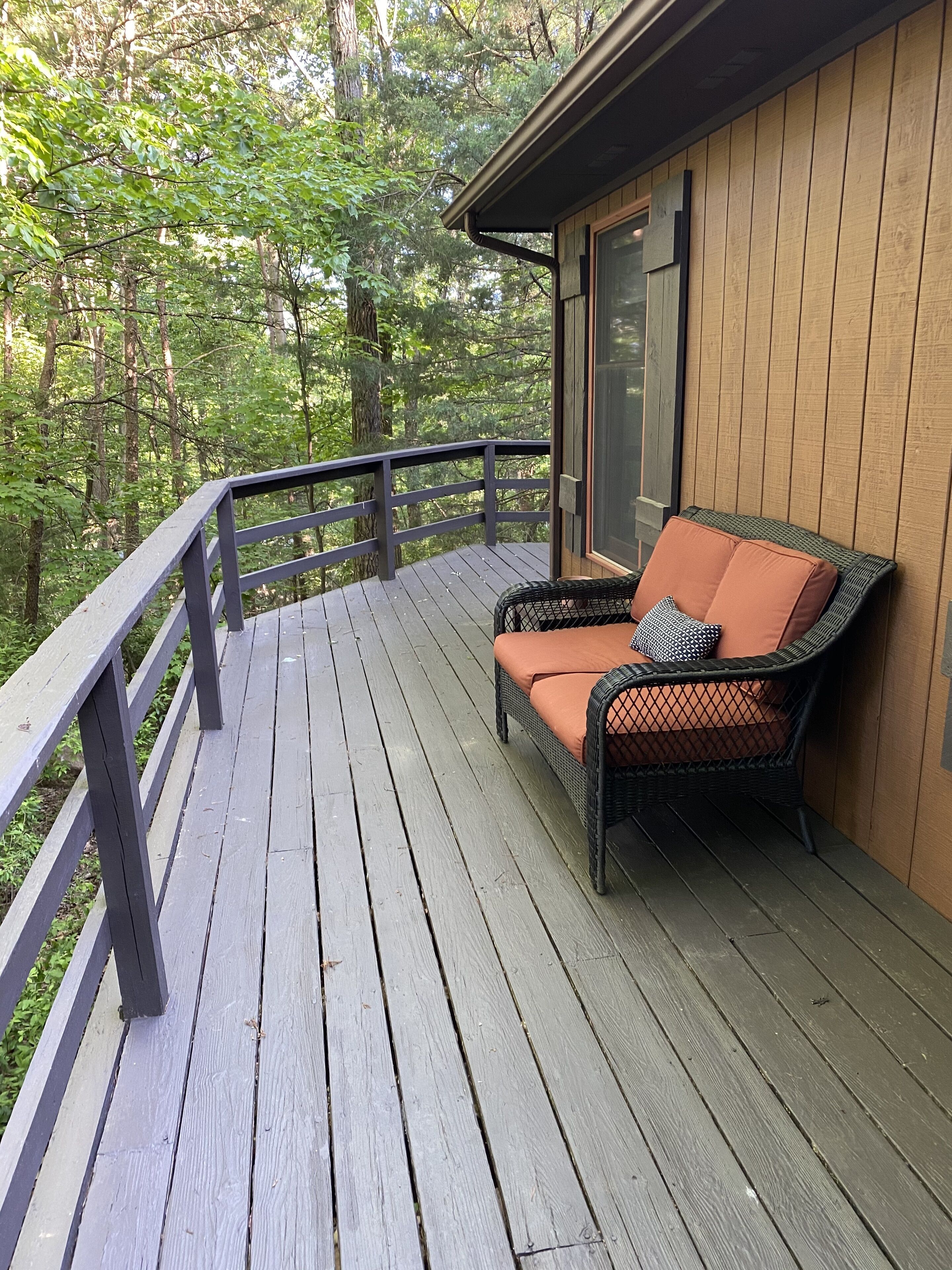 A wooden deck with a wicker chair and cushions, surrounded by a railing, overlooking a forested area.