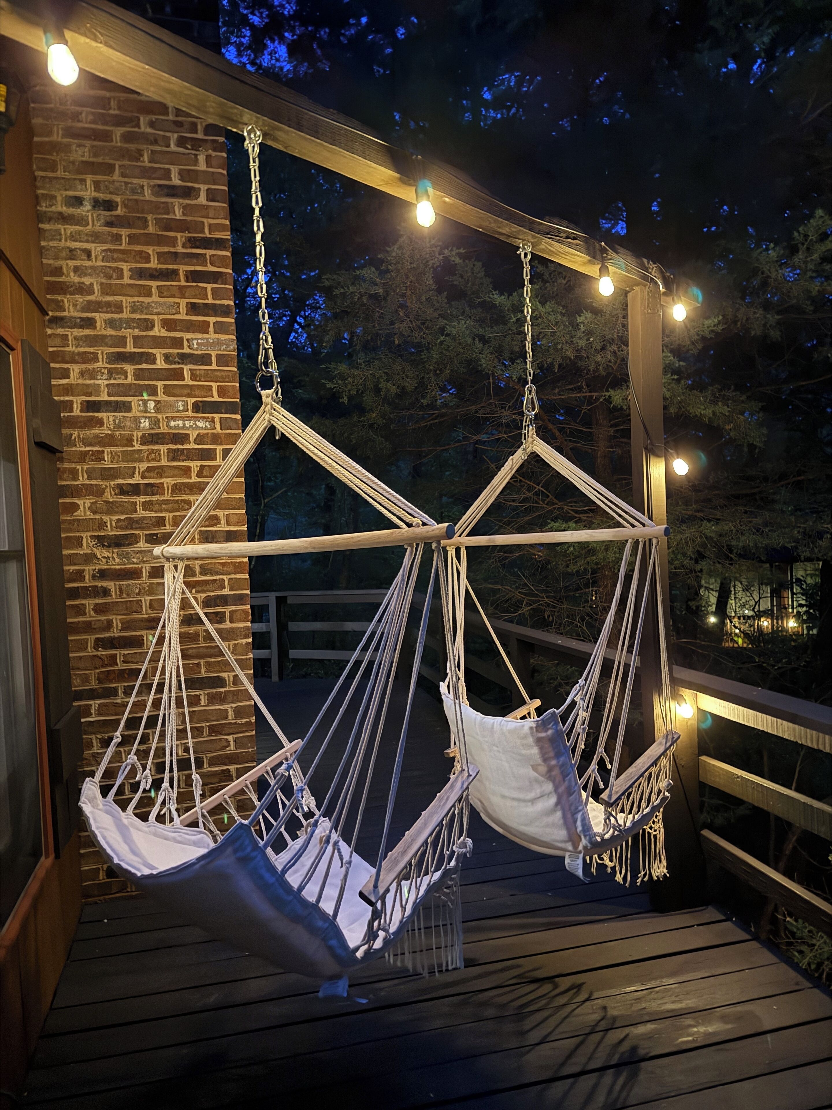 Una terraza de madera con dos hamacas blancas colgadas de un armazón, rodeada de luces colgantes.