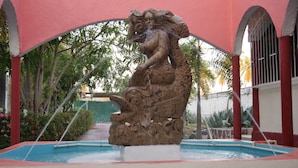 Eine Steinskulptur einer Fontäne in einem Innenhof mit Teich, umgeben von Bäumen und einem Gebäude mit roten und weißen Wänden.