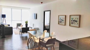 A modern living room with a glass dining table, comfortable seating, a large window, and a flat-screen TV mounted on the wall. - Two Beautiful 1 BR apts, 2 floors apart HS WiFi, full amenities! (Ciudad de México)