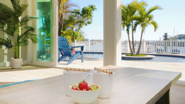 Una mesa con dos tazas blancas y un bol de fruta en un balcón con vistas a una piscina y palmeras.