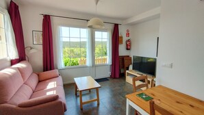 A living room with a pink sofa, a wooden table, a TV, and large windows with curtains. - Apartamentos Corona (Comillas)