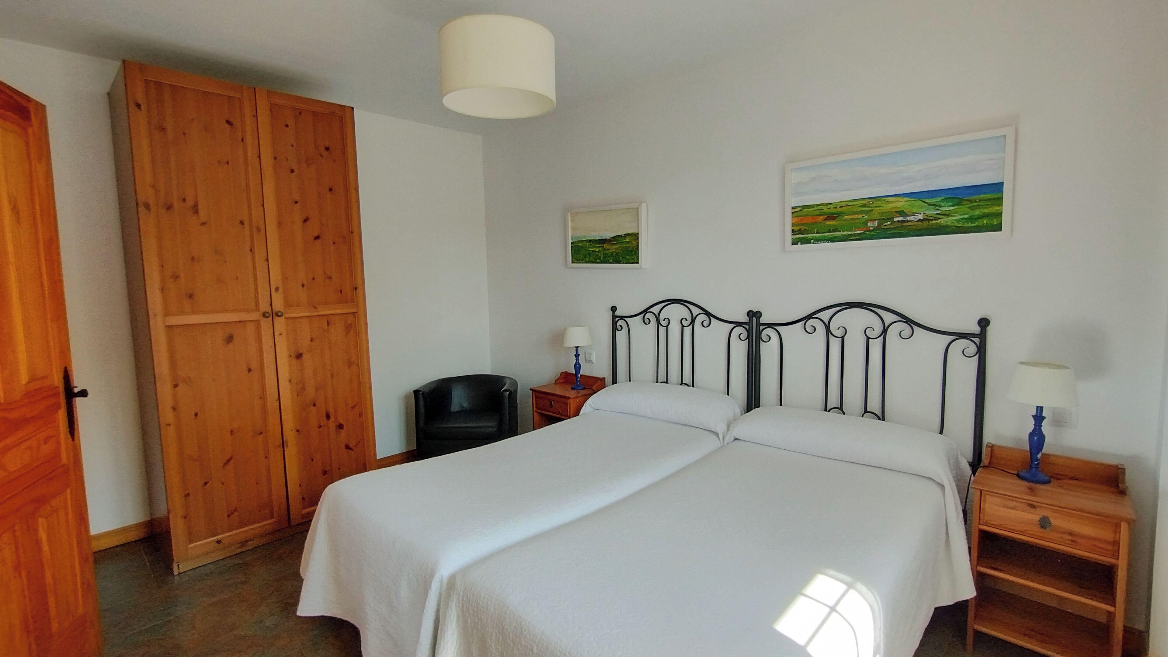 A bedroom with a double bed, wooden wardrobe, two bedside tables with lamps, and a landscape painting on the wall.