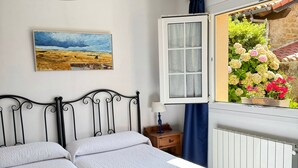 A bedroom with two beds, a hanging light fixture, a window with a view of flowers, and a painting on the wall. - Apartamentos Corona (Comillas)