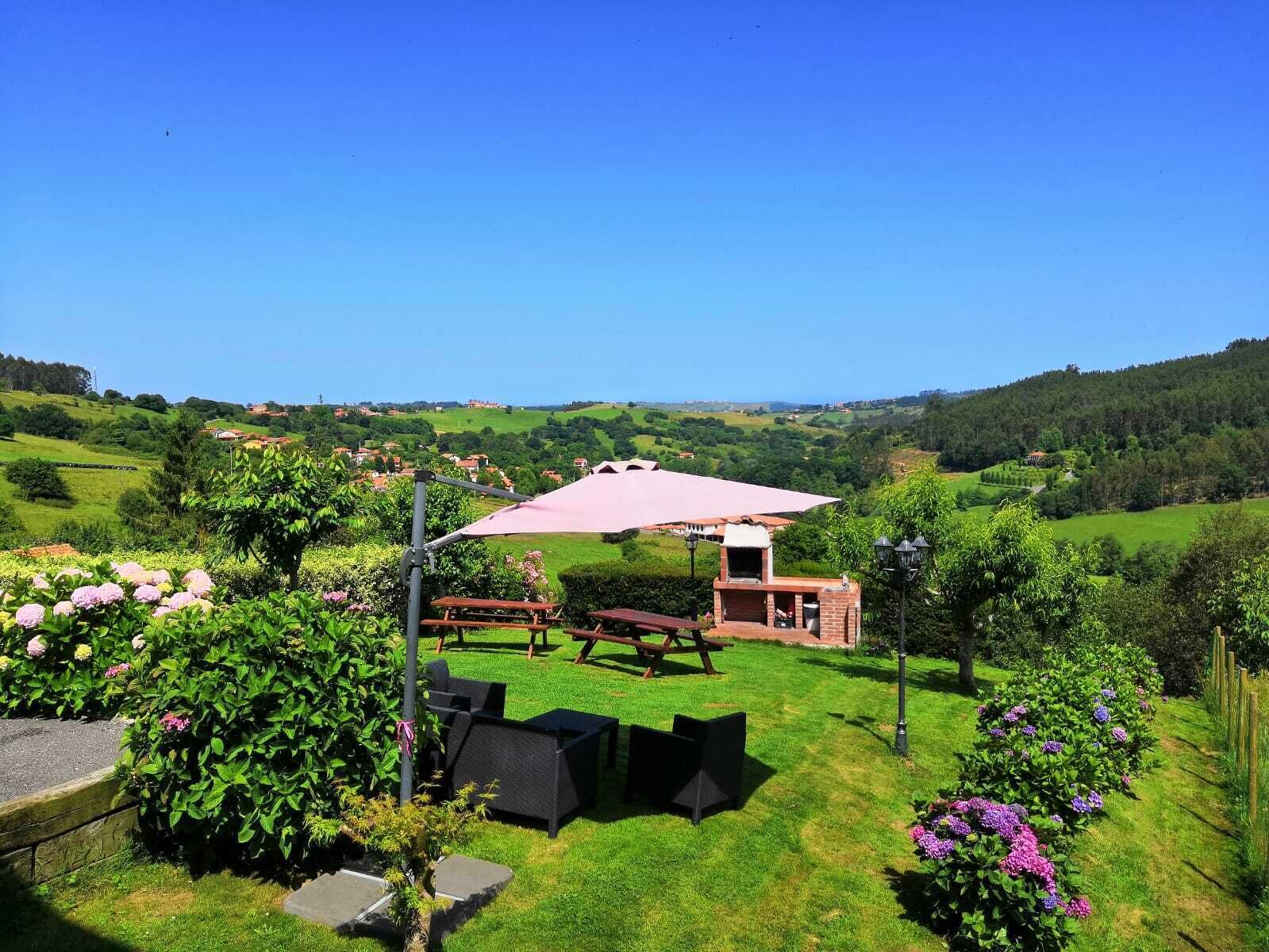 Un espacio al aire libre con césped, mesas de picnic y una parrilla, rodeado de abundante vegetación y una vista panorámica de colinas ondulantes.