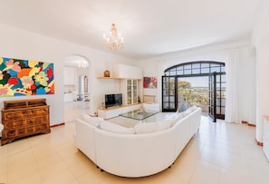 A spacious living room with a white sofa, a glass coffee table, and a TV. There's a large window with a view, a chandelier, and a colorful abstract painting on the wall.