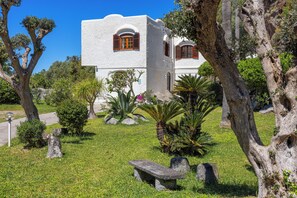 A white building with arched windows and a stone bench in a garden with trees and plants.