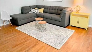 A living room with a grey sectional sofa, a patterned rug, two wooden side tables, a white chair, a yellow cabinet, and a framed picture on the wall.