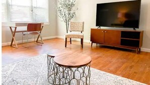 A living room with a wooden floor, a round coffee table, a television on a wooden cabinet, and a chair with a patterned cushion.