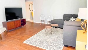 A modern living room with a flat-screen TV, grey sofa, two white chairs, a patterned rug, and a ceiling fan.