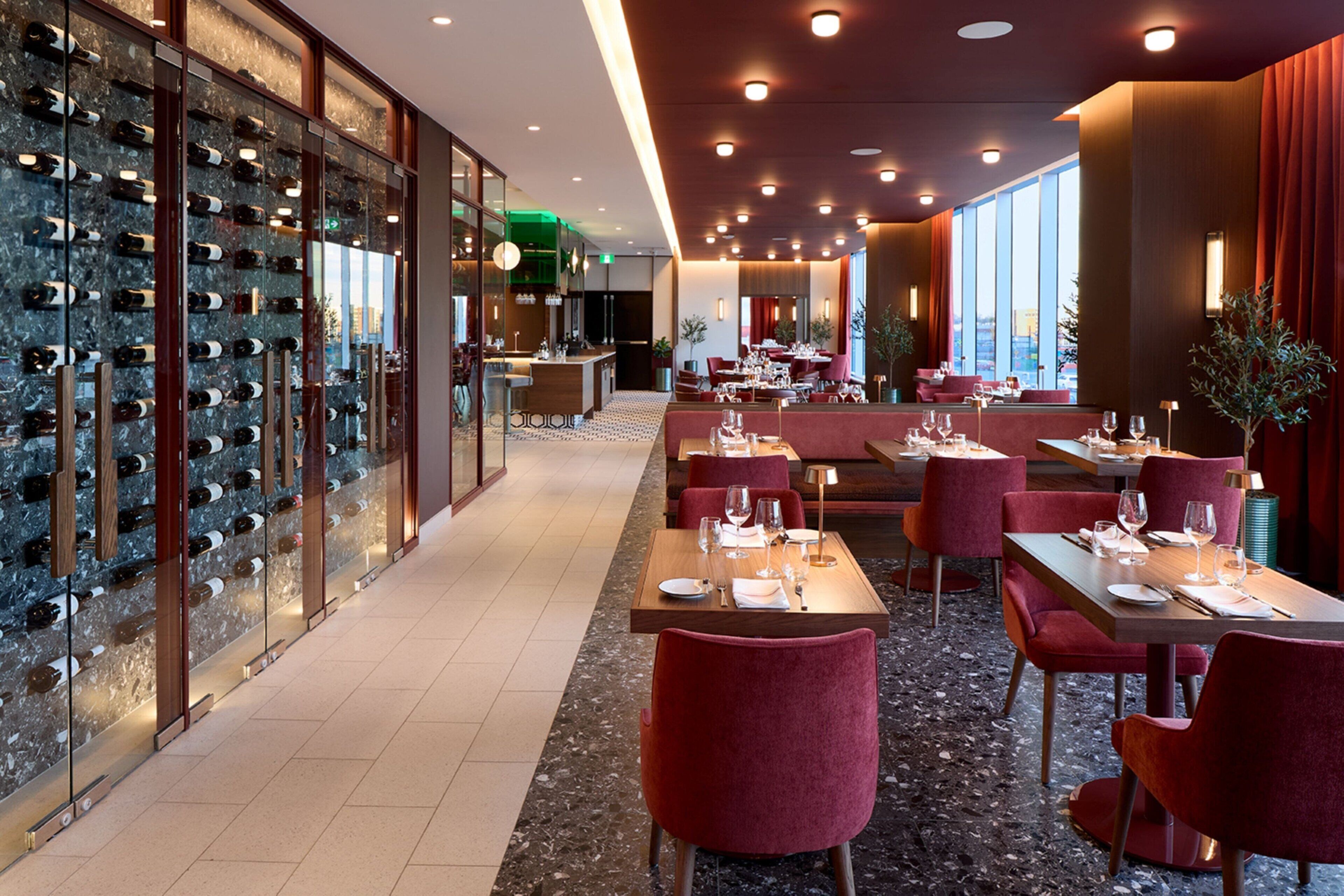 A restaurant with a wine wall, red upholstered chairs, and a bar area.