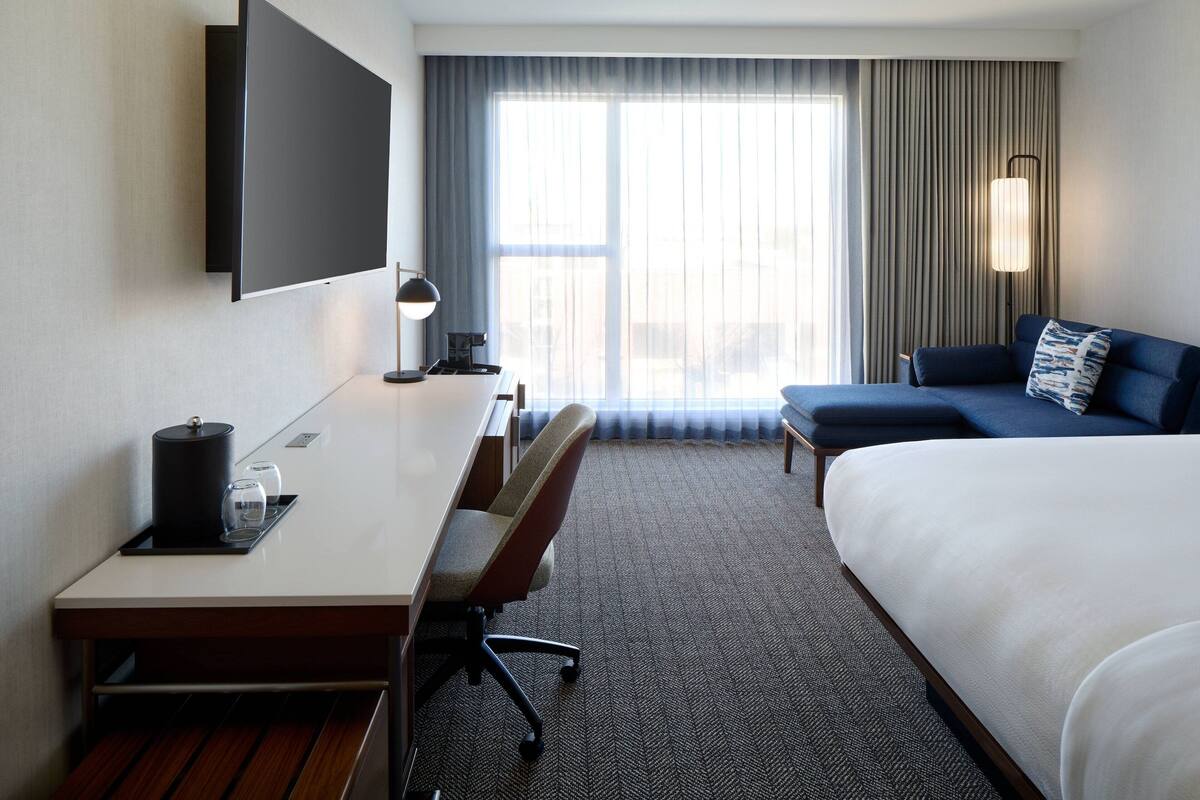 Una habitación de hotel con una cama, un escritorio con silla, un televisor montado en la pared y una ventana con cortinas.