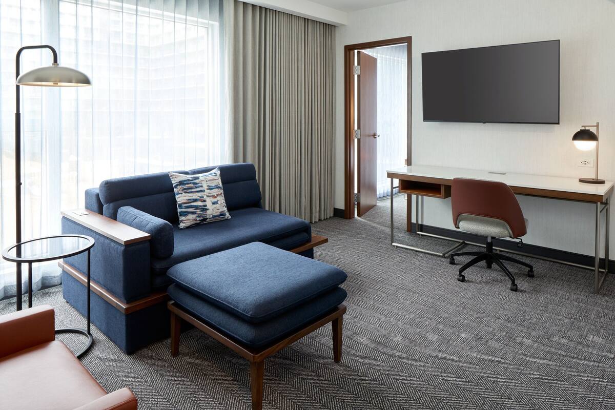 Una habitación de hotel con sofá, otoman, escritorio, silla, televisor y lámpara.