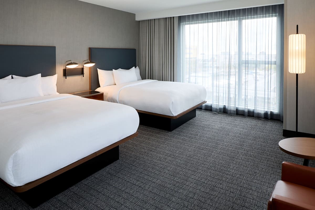 Una habitación de hotel con dos camas, un ventanal con cortinas, una mesa redonda y una lámpara.