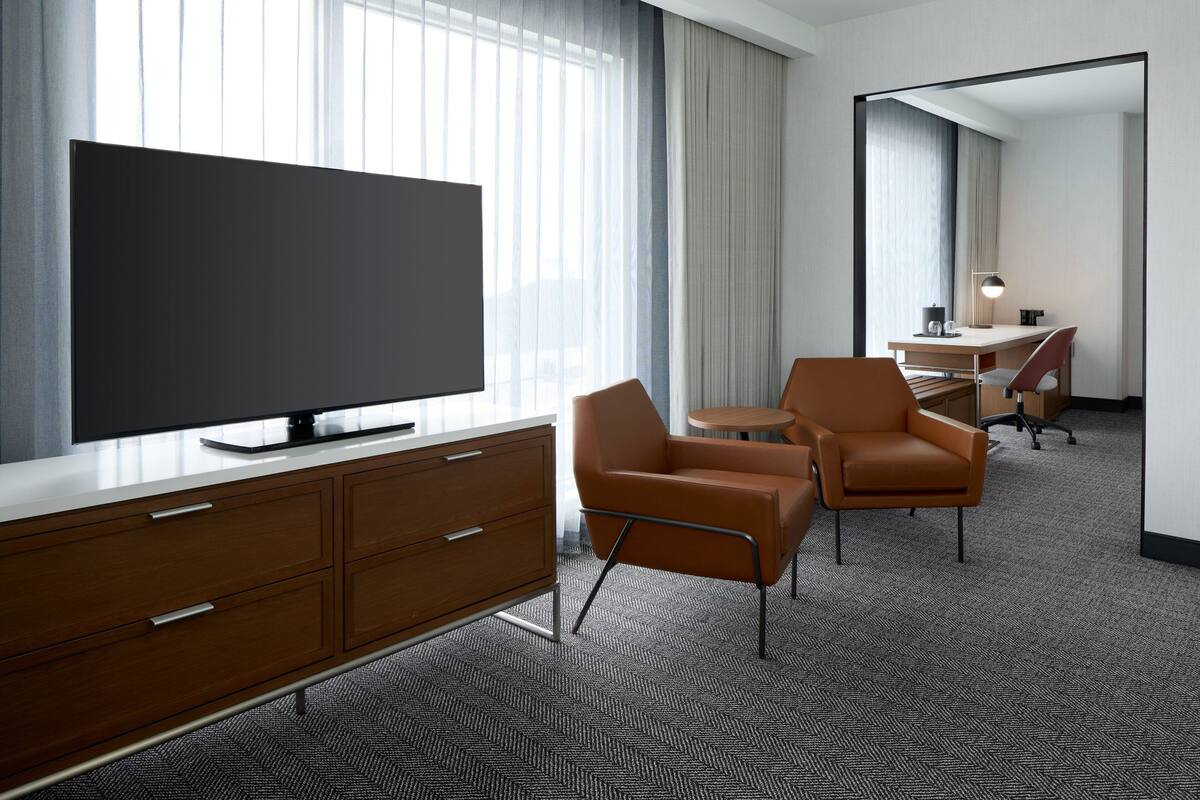 Una habitación de hotel moderna con un televisor de pantalla plana grande, un aparador de madera, dos sillones de cuero, una mesita redonda y un escritorio con una lámpara.