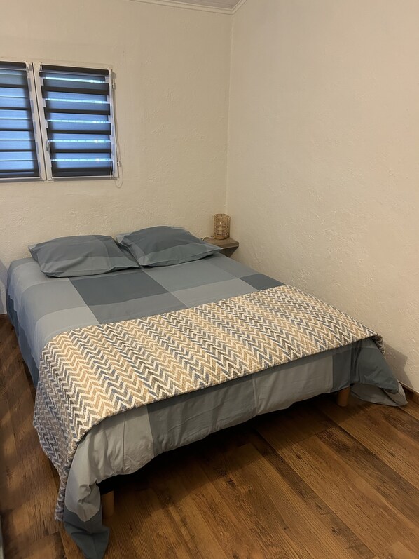 Ein Schlafzimmer mit einem Bett, einem Nachttisch, einem Fenster mit Jalousien und einem gewebten Korb auf dem Nachttisch.