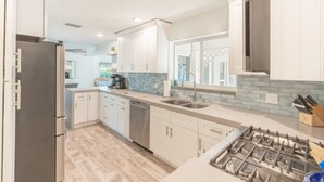 Una cocina moderna con armarios blancos, un refrigerador de acero inoxidable, lavavajillas, estufa a gas y una ventana con vista a follaje.
