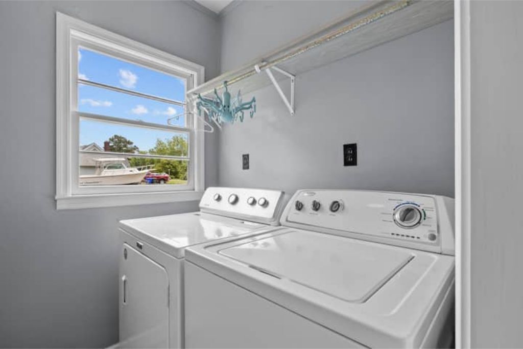 Uma lavanderia com duas máquinas de lavar e secar brancas, uma janela com vista para uma casa e um carro, e um lustre azul.