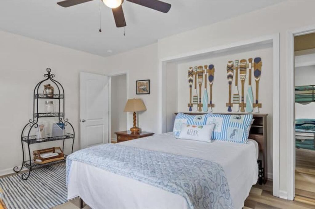 Une chambre à coucher avec un lit, un ventilateur de plafond, une étagère fixée au mur ornée d’objets décoratifs et une vue sur la mer, visible à travers une porte ouverte.