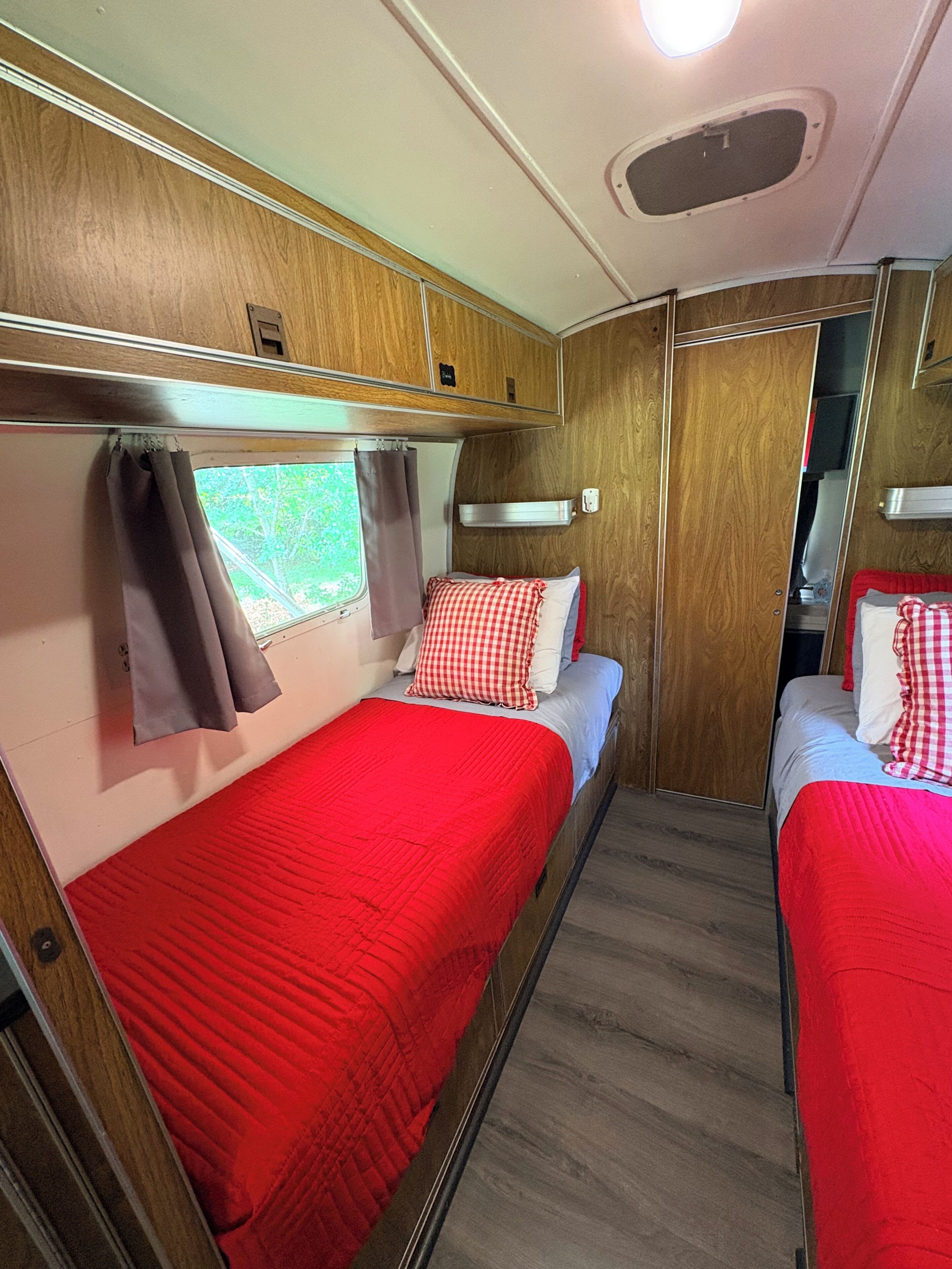 A compact RV interior with two beds, a window with curtains, and a small overhead light.