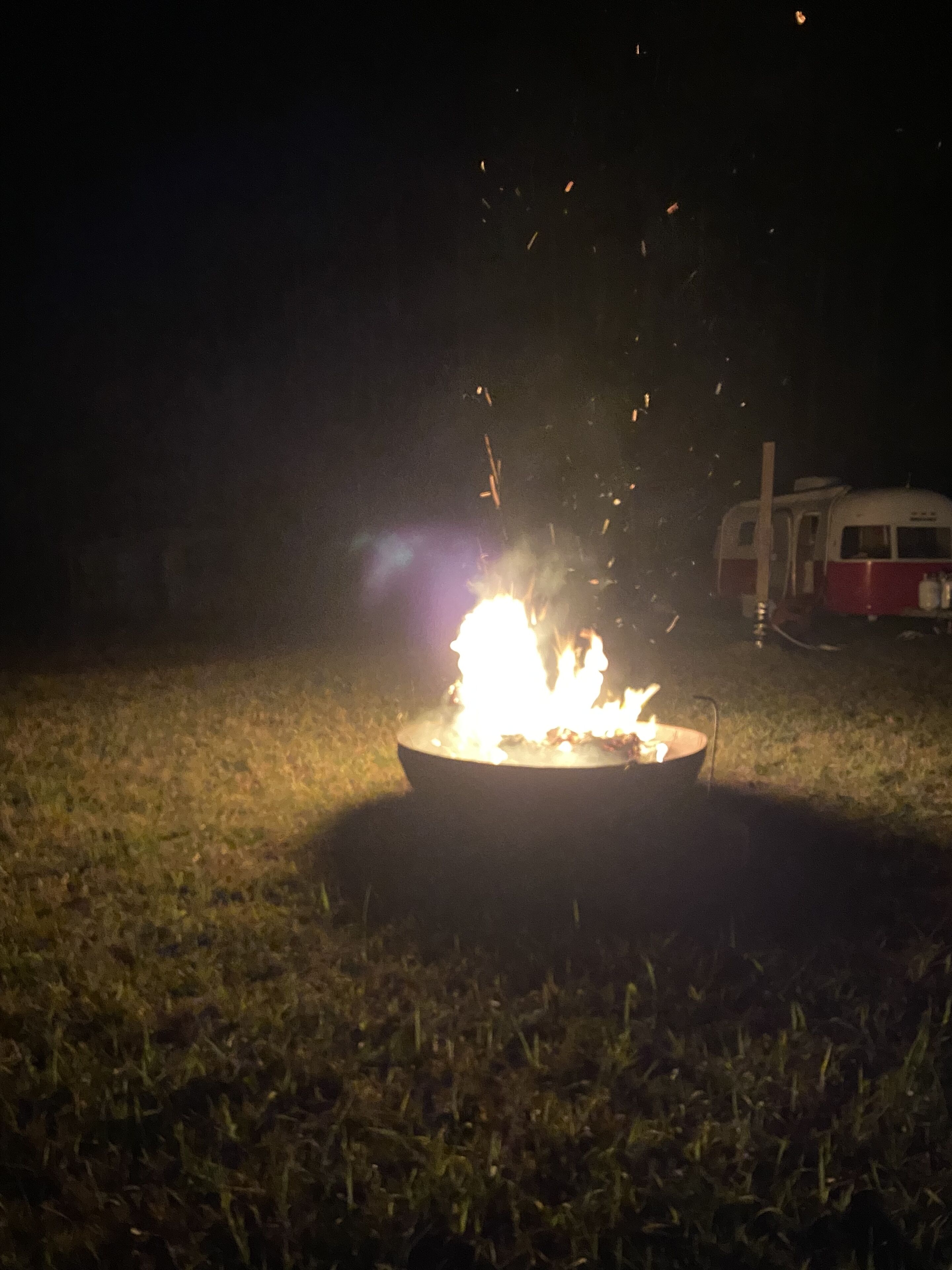 A campfire with sparks flying, a trailer in the background, and a grassy field.