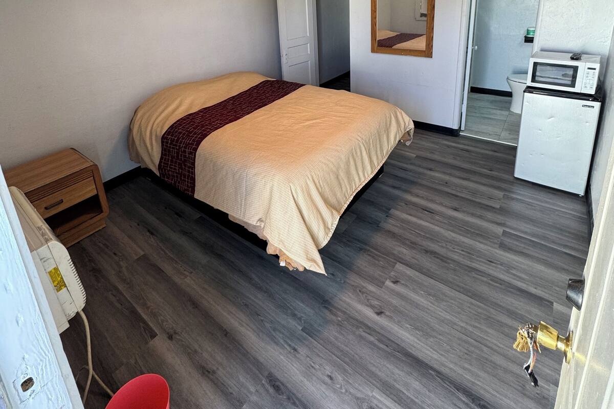 A bedroom with a bed, a small wooden nightstand, a red bucket, a white refrigerator, and a microwave.